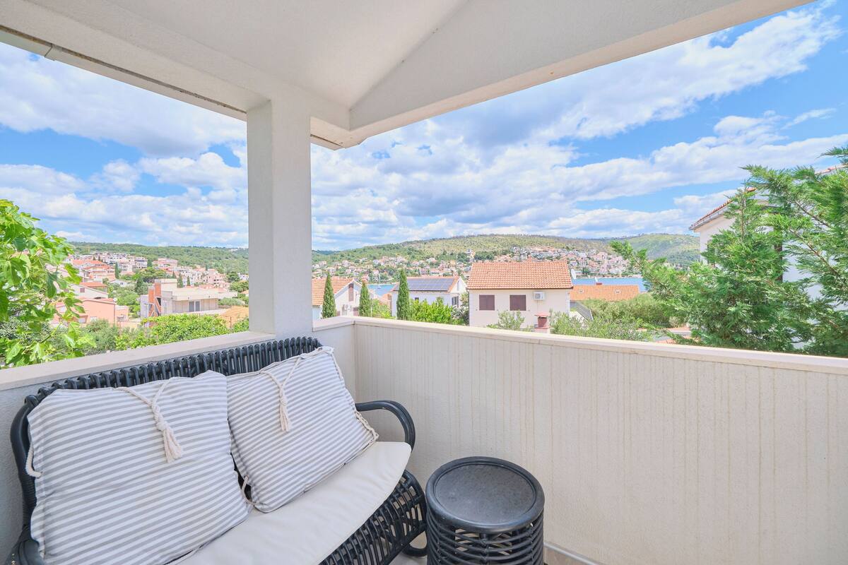 Ein Balkon mit einer Rattan-Couch und einem kleinen runden Tisch, mit Blick auf eine Stadt mit roten Dächern und grünen Hügeln.
