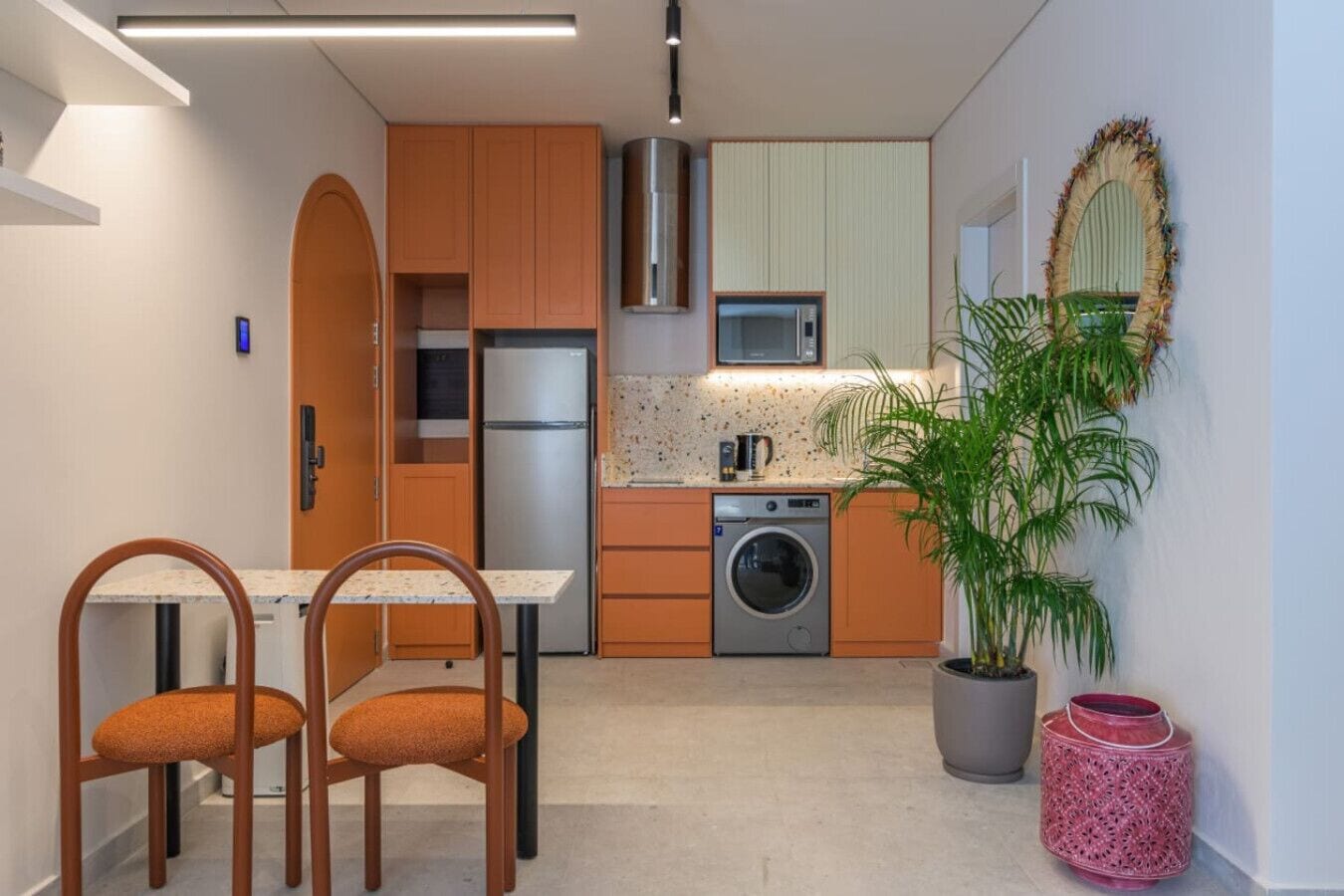 A compact kitchen with wooden cabinets, a built-in washing machine, a microwave, and a dining area with two chairs.