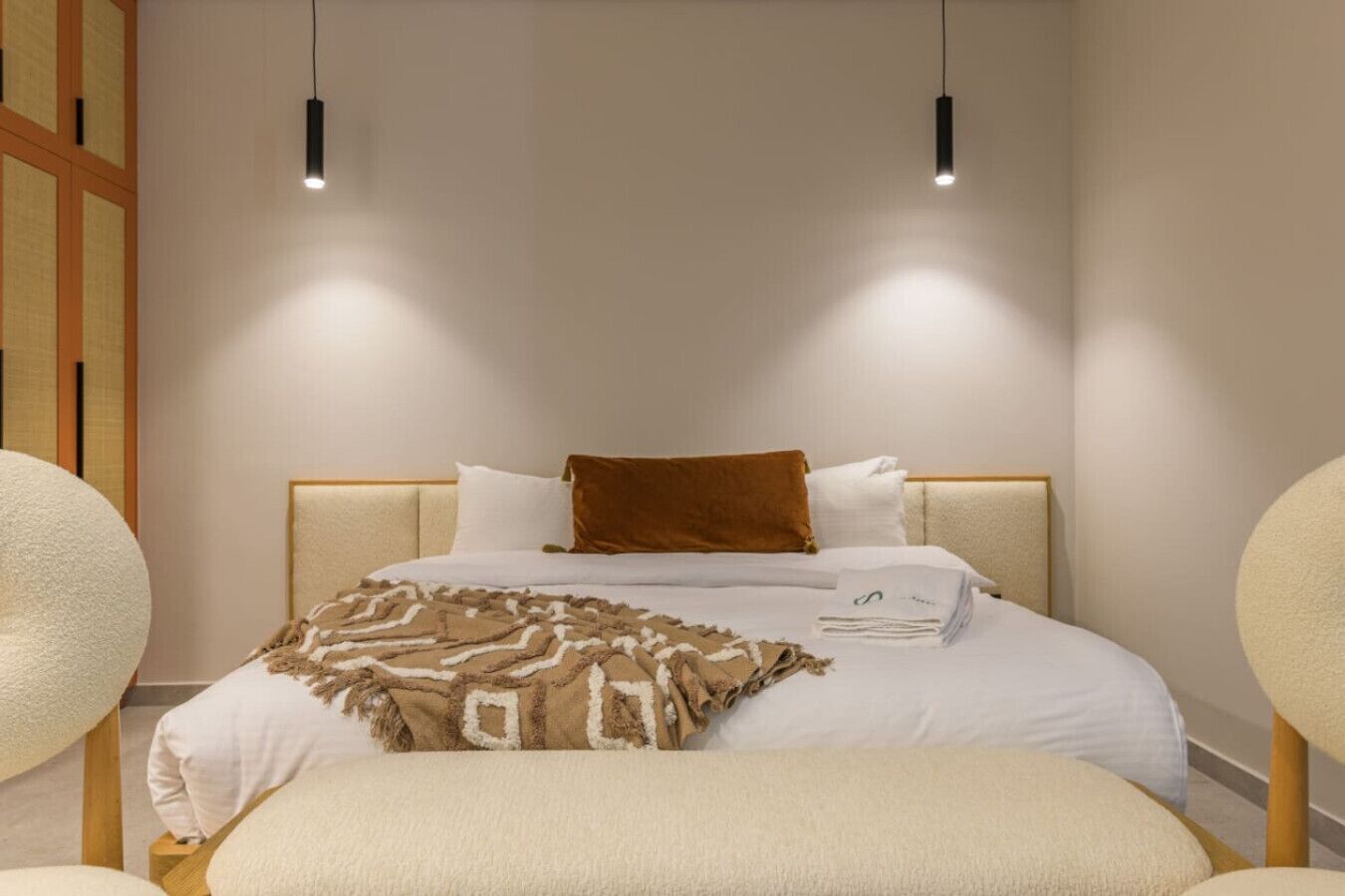 A neatly made bed with a brown throw blanket, white pillows, and a folded towel. There are two pendant lights above the bed.
