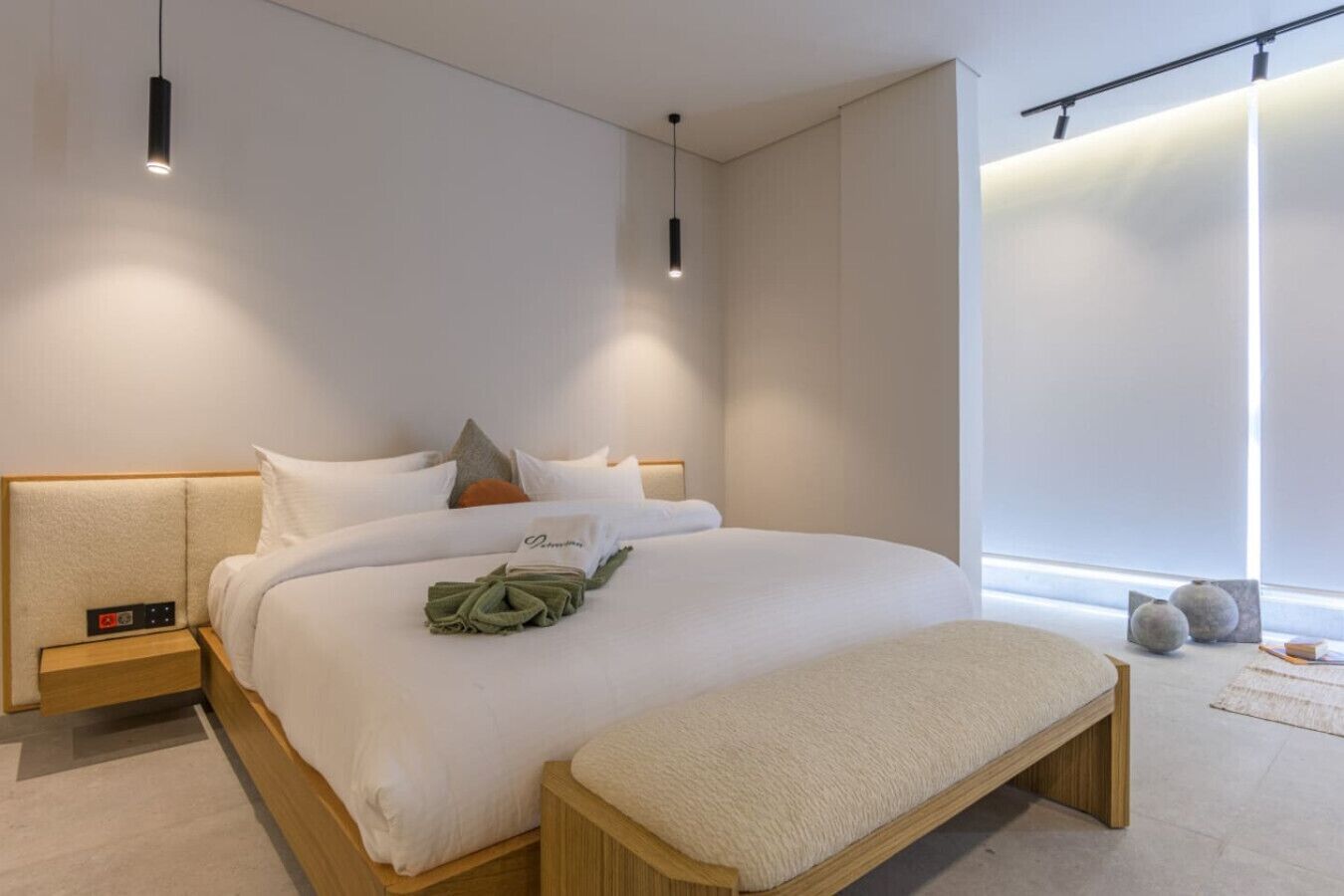 A modern bedroom with a large bed, bedside tables, a bench, and a window with blinds.