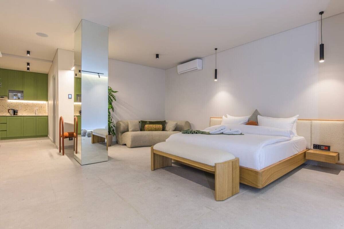 A modern hotel room with a large bed, a sitting area with a sofa, a small table, and a green kitchen area.