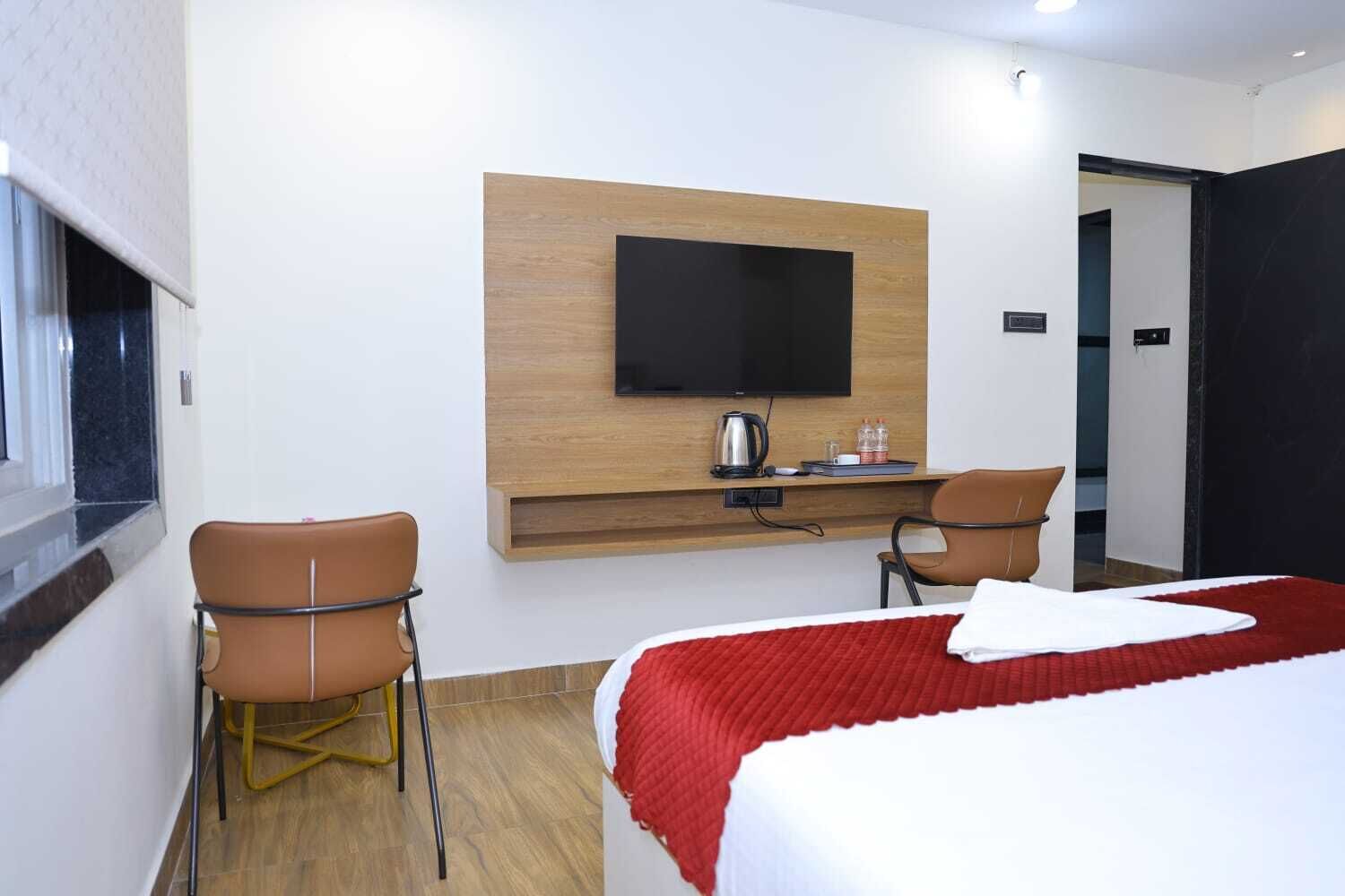 A hotel room with a bed, two chairs, a TV mounted on a wooden panel, a coffee maker, and a red blanket.