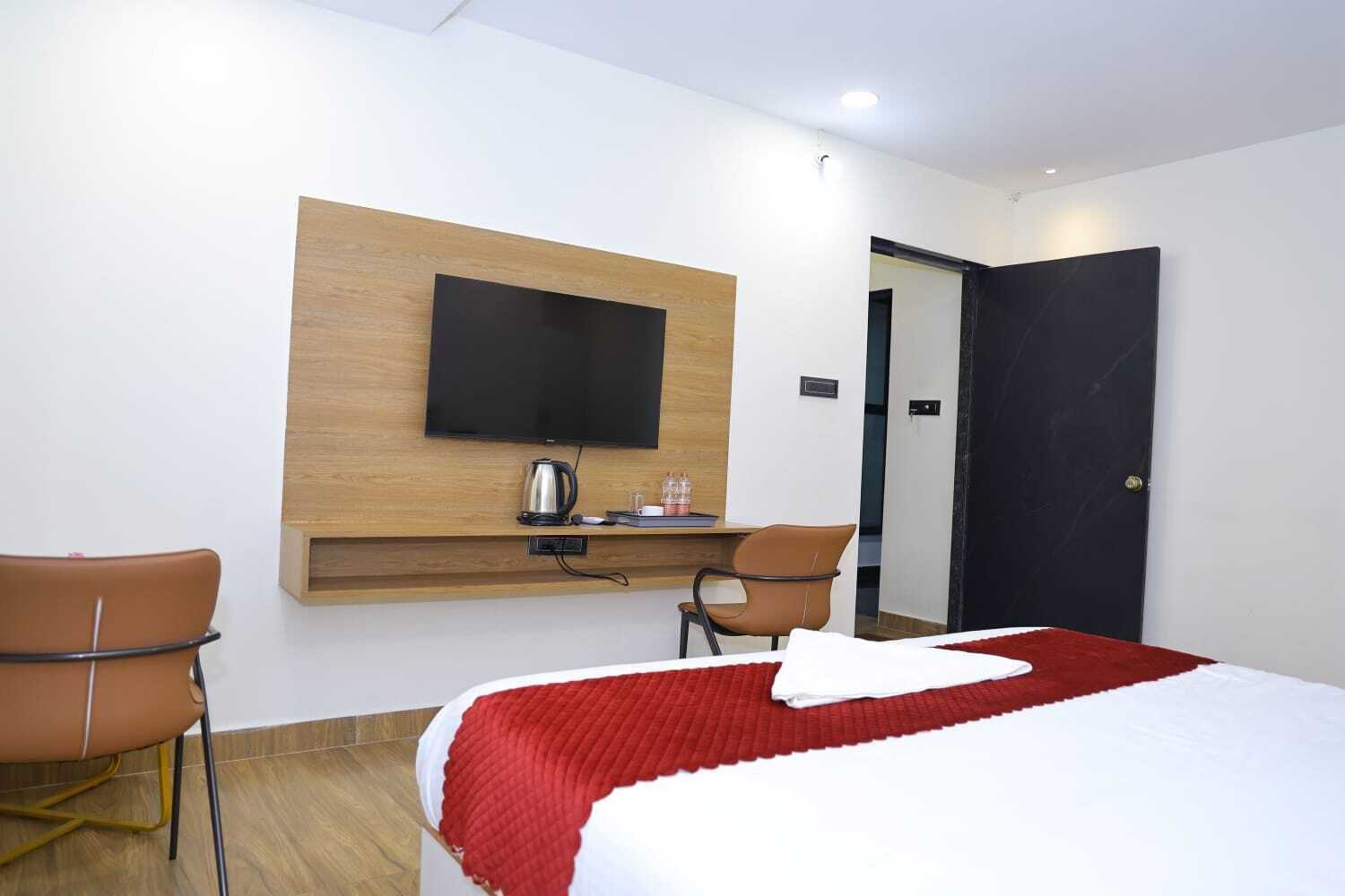 Hotelkamer met een bed, een televisie bevestigd aan een houten paneel, een bureau met een waterkoker, twee stoelen en een deur.