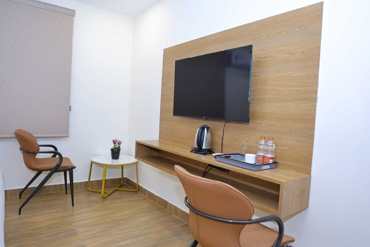 A small, modern room with a wooden wall-mounted TV, two brown chairs, a small round table with a potted plant, and a shelf with a kettle, glasses, and bottles.