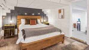 Ein modernes Schlafzimmer mit einem hölzernen Bett, einem Kopfteil mit integrierter Beleuchtung, Nachttischen und Blick in einen weiteren Raum.