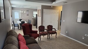 Un salón con sofá, sillones y televisor. Se observa un comedor con mesa y sillas al fondo.