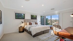 A bedroom with a large bed, a desk, and a view of a cityscape through large windows. - Metawise Luxe 6 Bed Ocean View Family Home in Seaforth (Sydney)