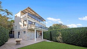 A modern two-story house with a glass balcony, stone facade, and a well-maintained lawn. - Metawise Luxe 6 Bed Ocean View Family Home in Seaforth (Sydney)