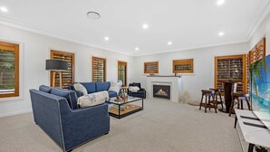 A spacious living room with a blue sofa, a fireplace, and a large TV screen. - Metawise Luxe 6 Bed Ocean View Family Home in Seaforth (Sydney)