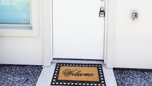 A white door with a 'D' marking, a doormat with 'Welcome' written on it, a blue window with blinds, and a wall-mounted light fixture. - Apartment With 3 Bedrooms 40 Minutes From South Padre (Weslaco)
