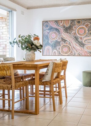 A dining area with a wooden table and wicker chairs, a vase with flowers, and a large abstract painting on the wall.