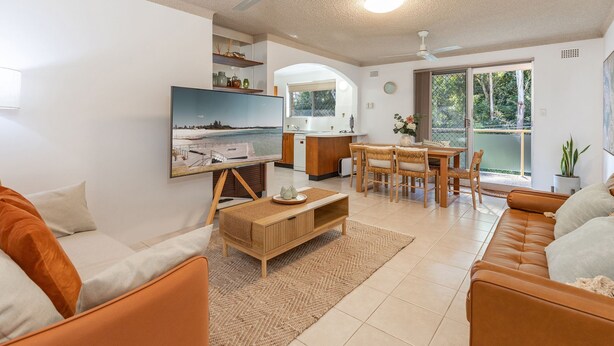 Una sala de estar moderna con sofá, mesa de centro y televisor. Hay una zona de cocina con mesa y sillas. La habitación tiene un ventanal grande y un ventilador de techo.