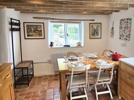 Un comedor con una mesa de madera dispuesta para cuatro personas, rodeada de sillas blancas. Hay una ventana que da a vistas de follaje y una maceta roja sobre la mesa.