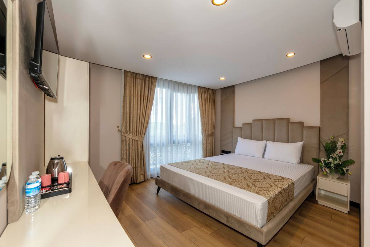 Ein Hotelzimmer mit einem großen Bett, einem Schreibtisch mit Wasserflasche und Kaffeemaschine, einem an der Wand befestigten Fernseher und einem Fenster mit Vorhängen.