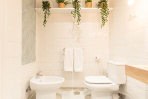 Ein Badezimmer mit weißen Fliesen, einem Waschbecken, einer Toilette und zwei Blumentöpfen auf Regalen.