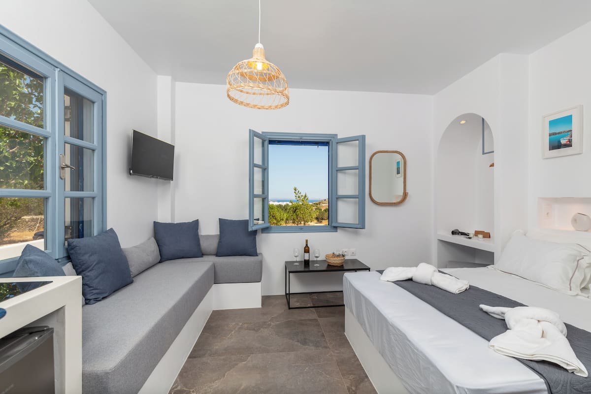 A modern hotel room with a sofa, a bed, a small table with a bottle and glasses, a TV, and a window with an open shutter.