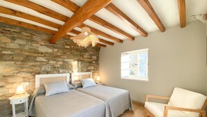 Une chambre avec deux lits, un mur en pierre, des poutres en bois, un luminaire suspendu, une fenêtre et une chaise en bois.