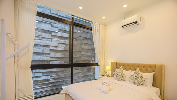 Habitación de hotel con un ventanal grande, cama con ropa de cama blanca, dos almohadones decorativos, mesita de noche con lámpara y un equipo de aire acondicionado en la pared.