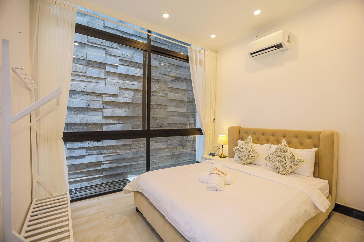 A hotel room with a large window, a bed with white bedding, two decorative pillows, a bedside table with a lamp, and an air conditioning unit on the wall.
