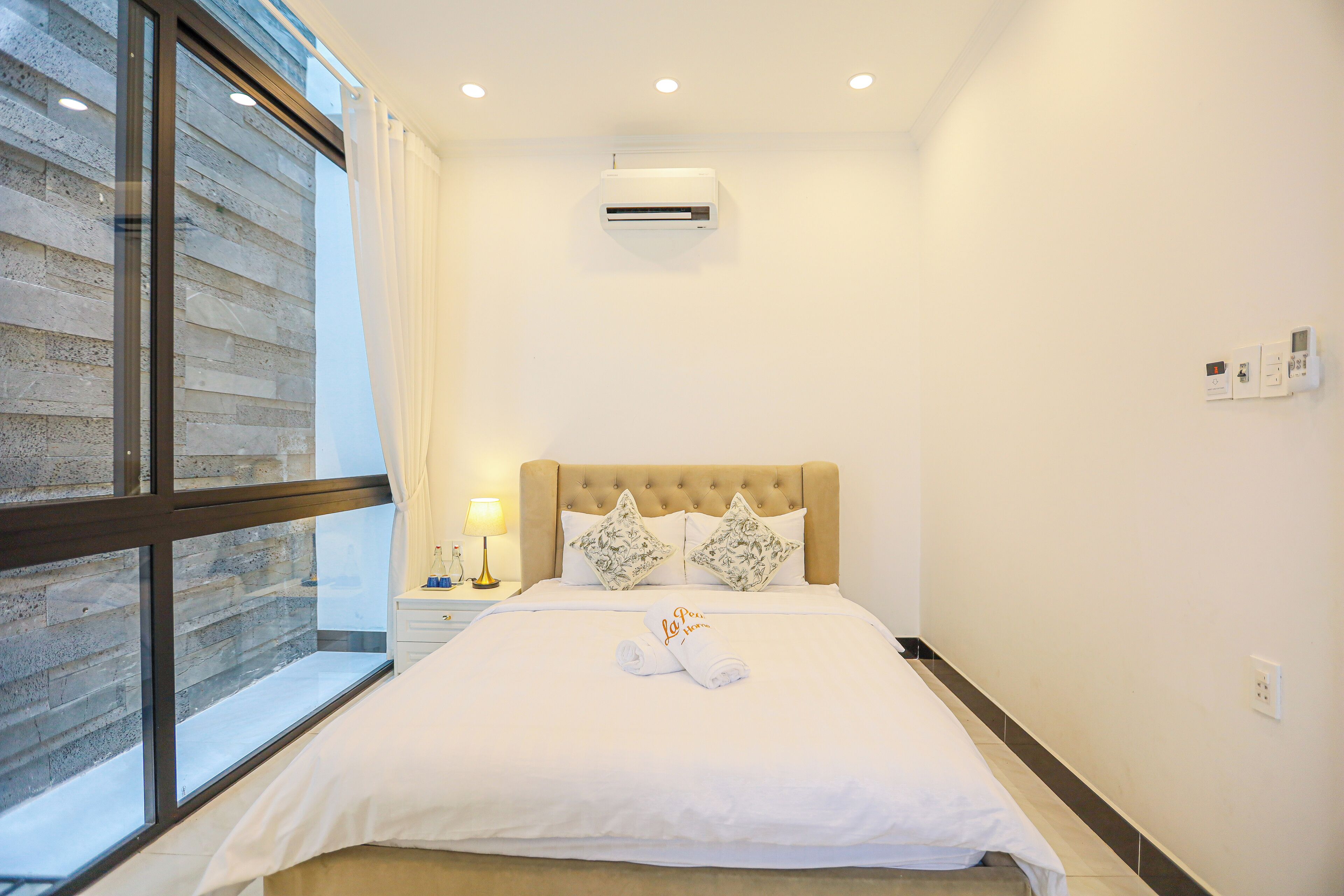 A hotel room with a large bed, a window with a view, an air conditioning unit, and a nightstand with a lamp.