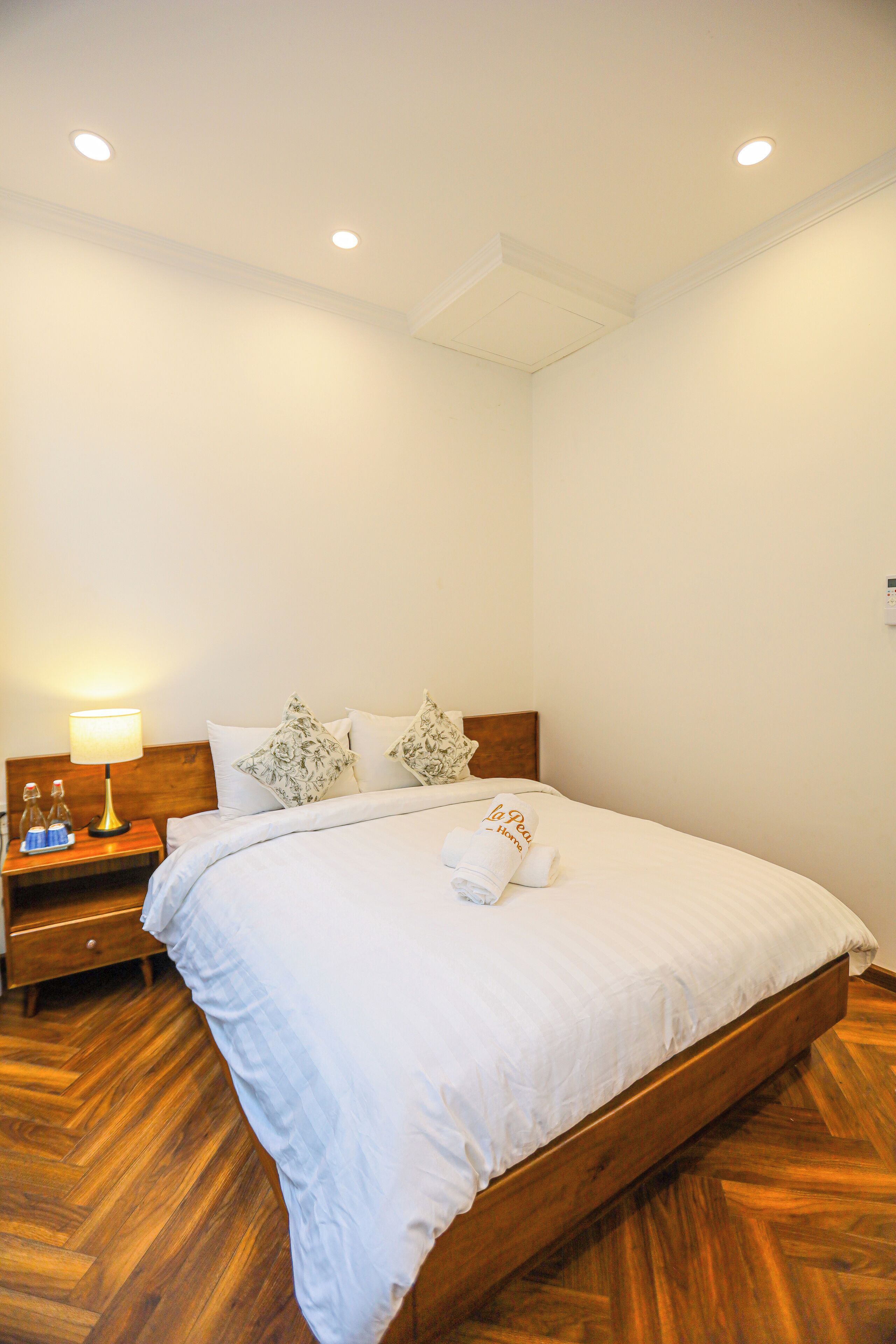 A neatly made bed with white linens and a rolled towel, a wooden nightstand with a lamp, and a wooden floor.