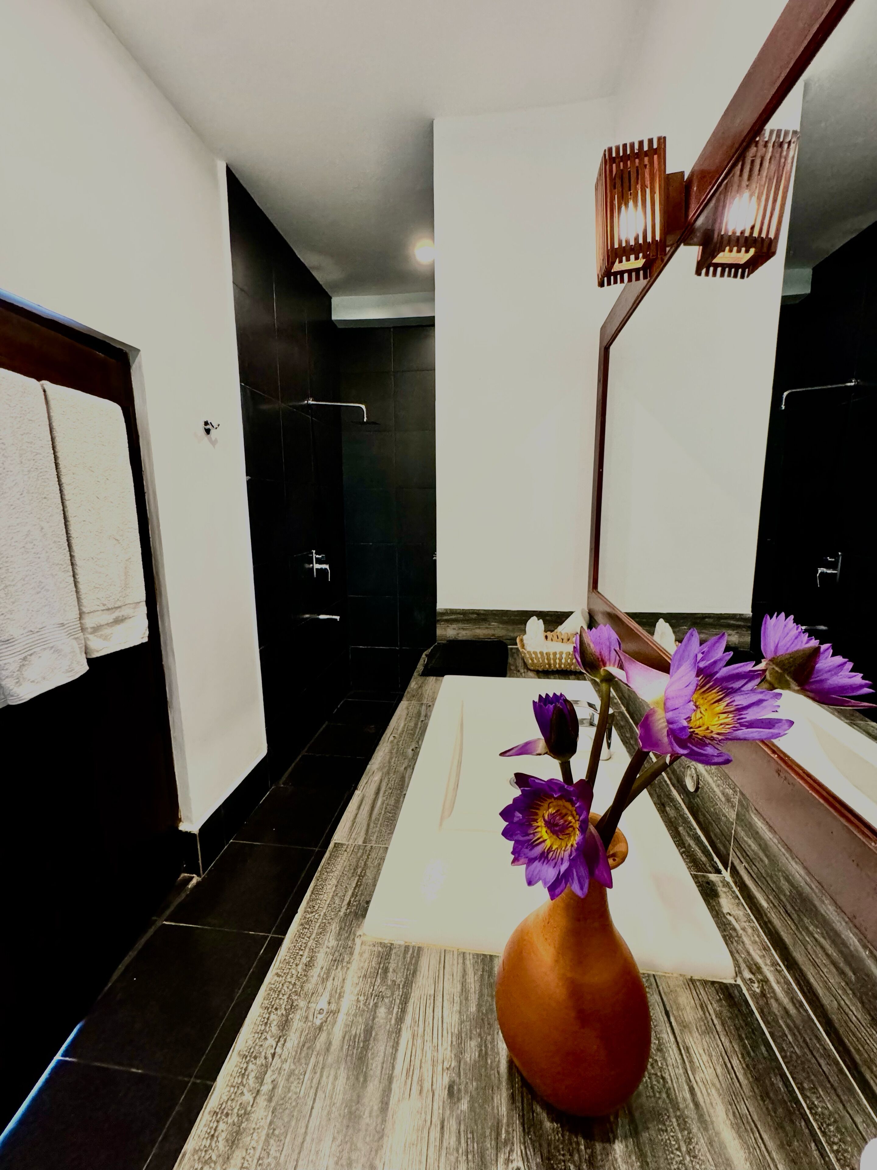 Um banheiro com piso de madeira, uma toalha branca pendurada em uma porta, um espelho e um vaso com flores roxas.