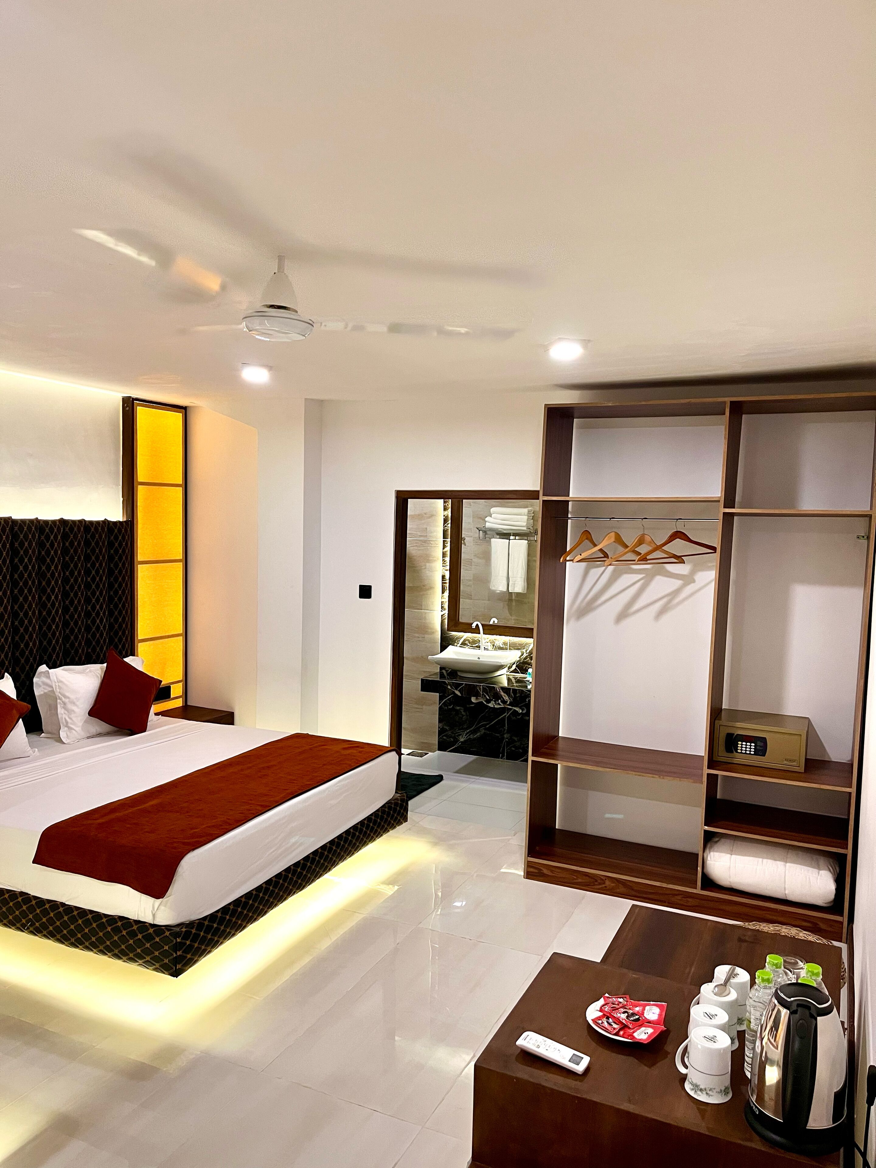 A modern hotel room with a neatly made bed, a bedside table with a remote control, a coffee table with breakfast items, and a walk-in closet with hanging clothes.
