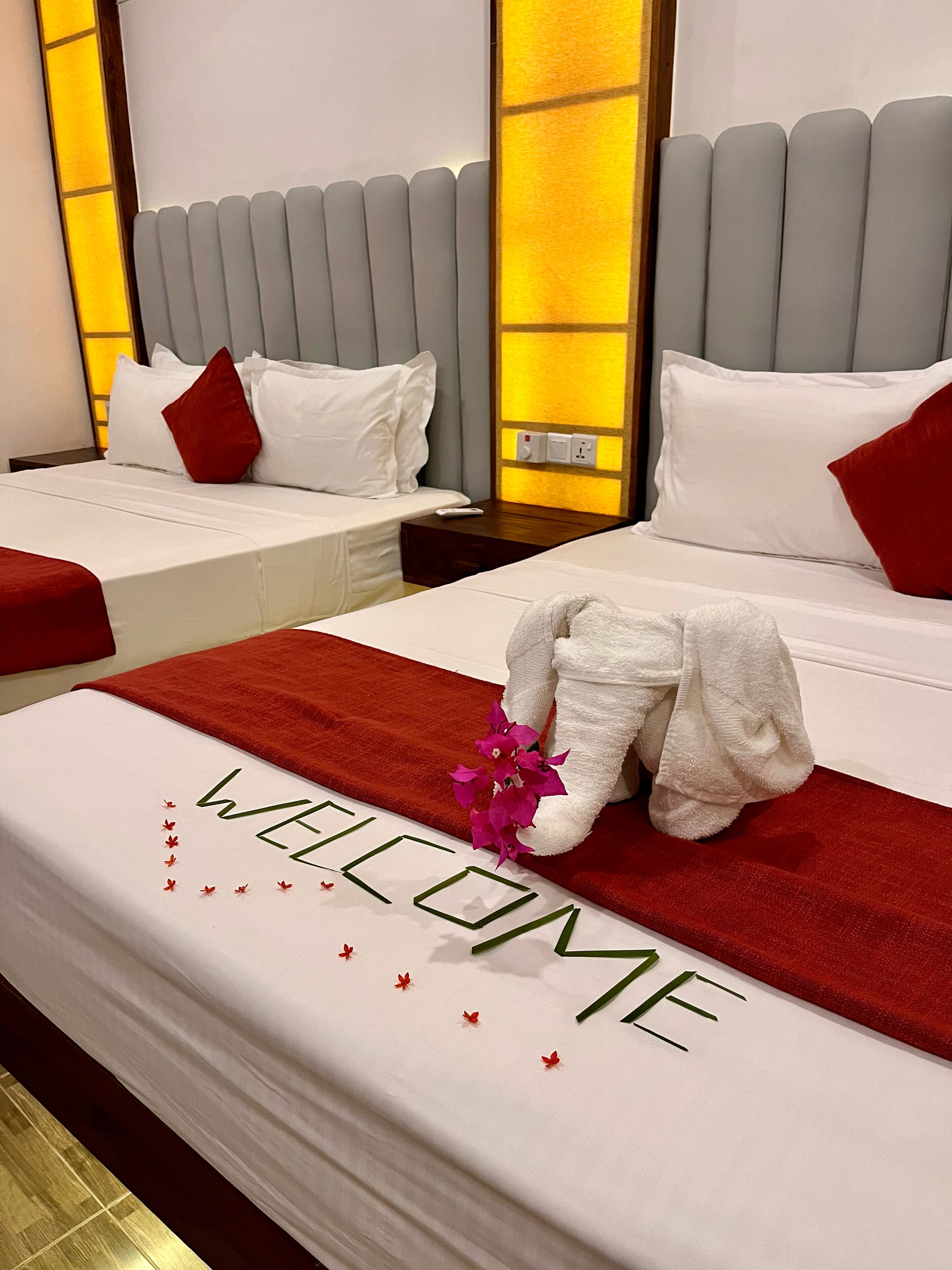 Une chambre d’hôtel avec deux lits, chacun agrémenté de draps blancs et d’oreillers rouges. Une serviette ornée d’une fleur est posée sur l’un des lits, et le mot « BIENVENUE » est brodé sur la couverture.