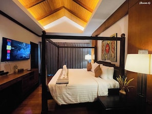 A hotel room with a four-poster bed, a flat-screen TV mounted on the wall, a bedside table with a lamp, and a decorative wall piece. - Kiri Anda Villa Phuket (Rawai)