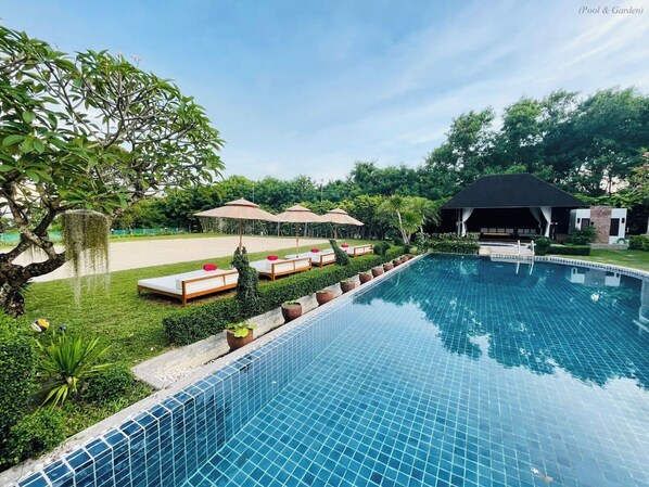 A swimming pool with lounge chairs and umbrellas, surrounded by greenery and a building in the background. - Kiri Anda Villa Phuket (Rawai)