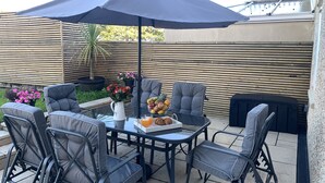 Une terrasse avec une table, des chaises, un vase de fleurs et un panier de fruits.