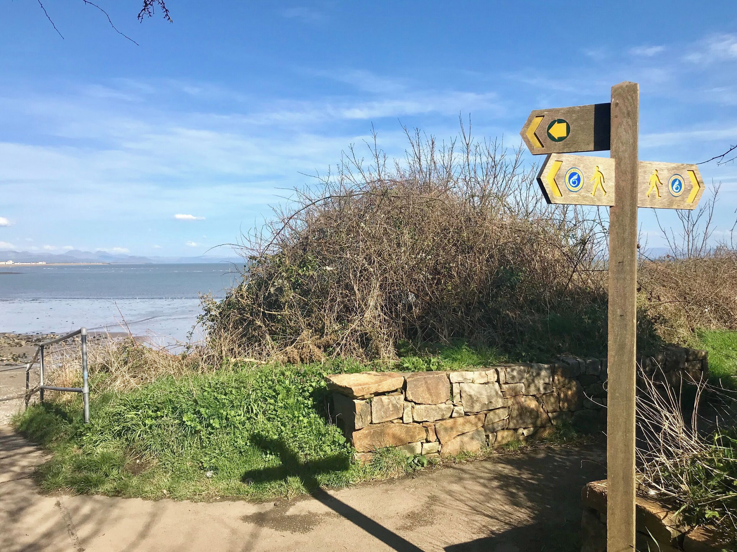 A wooden signpost with directional arrows, a stone wall, and a view of the sea.