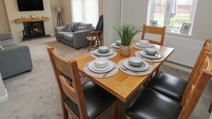 Un coin repas avec une table en bois dressée pour quatre personnes, entourée de chaises en bois. On aperçoit une cheminée au fond, et un téléviseur est fixé au mur.