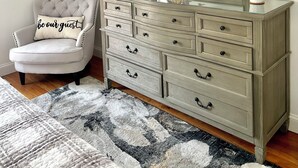 Una habitación con un sillón blanco, un aparador con varios cajones, un espejo, una lámpara y un estante pequeño con objetos decorativos.