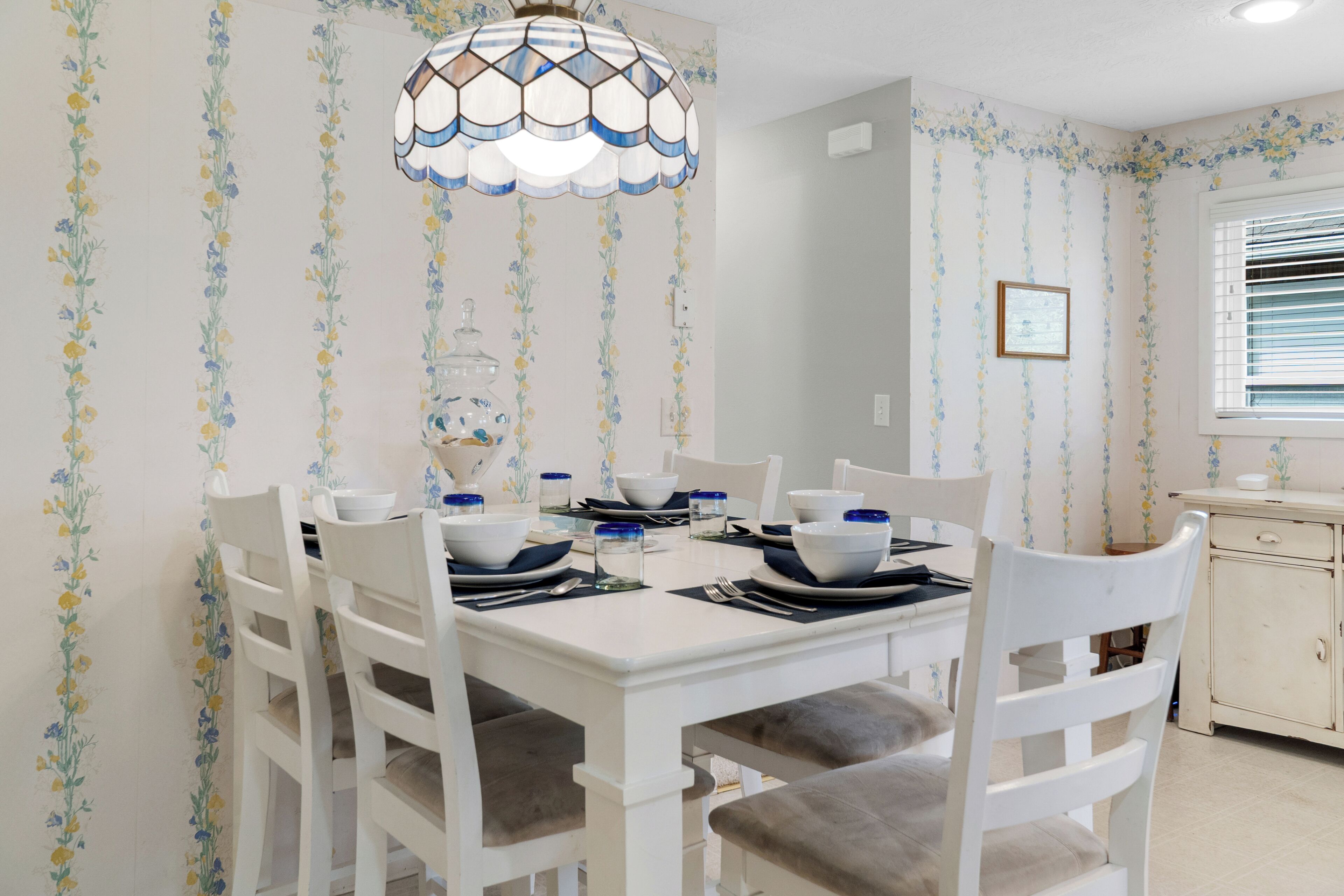 A dining area with a white table set for four, surrounded by white chairs with grey cushions. A decorative glass pendant light hangs above the table.