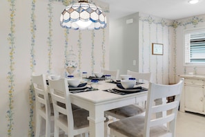 A dining area with a white table set for four, surrounded by white chairs with grey cushions. A decorative glass pendant light hangs above the table.