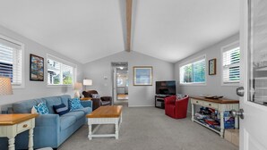 Ein Wohnzimmer mit einem blauen Sofa, einem Holztisch und einem roten Hocker. Ein Fernseher und ein Bücherregal mit Spielen sind ebenfalls vorhanden.