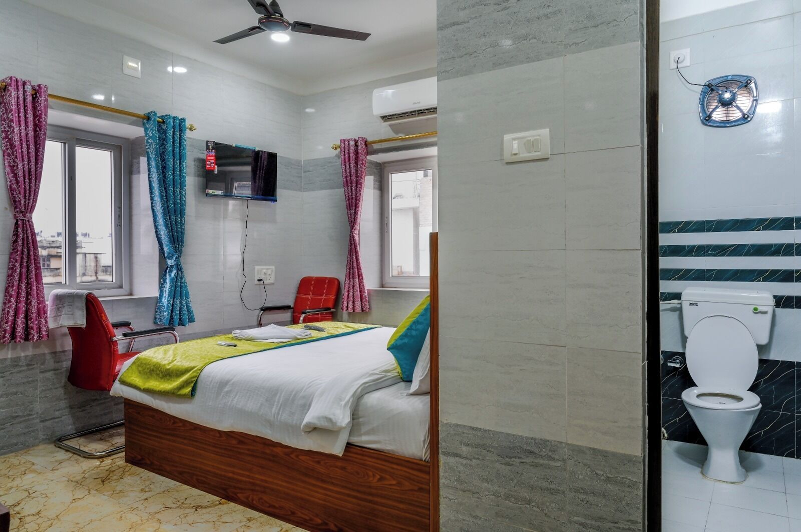 Habitación de hotel con cama, una silla roja, televisión y baño con inodoro y ventilador.