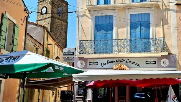 Une vue de rue avec un bâtiment portant l’inscription « Les Trois Petits Cochons » et un beffroi.
