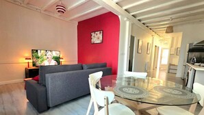 Un salon moderne avec un canapé gris, une table à manger en verre entourée de chaises, un mur d’accent rouge et une cuisine en arrière-plan.