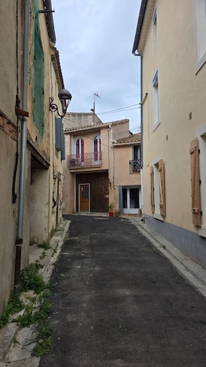 A narrow street with buildings on either side, a visible door, and a small balcony with a railing.