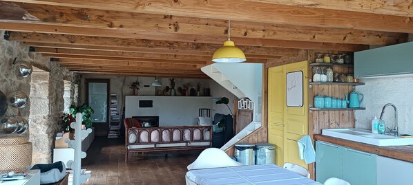 A rustic interior with a wooden ceiling, a yellow pendant light, a sofa, a staircase, and a kitchen area with jars and a sink.
