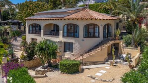 A Mediterranean-style villa with a terracotta-tiled roof, a stone facade, and a central staircase leading to a balcony.