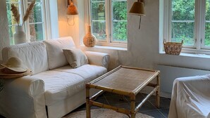 Ein gemütliches Zimmer mit einem weißen Sofa, einem gewobenen Couchtisch und großen Fenstern mit Blick ins Grüne.