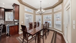 A spacious dining area with a large table and chairs, a kitchen with dark wood cabinets, and large windows with blinds. - Big Family & Group-Friendly 9BR Home in NW Calgary (Calgary)