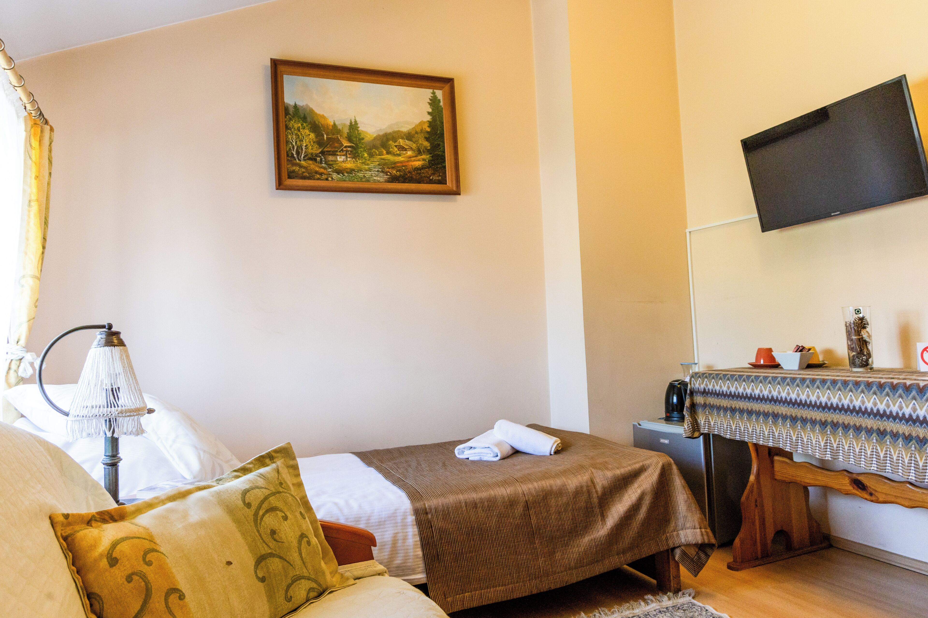 Una camera d'albergo con un letto, un tavolino con tovaglia decorativa, una televisione fissata al muro e un quadro sopra il letto.