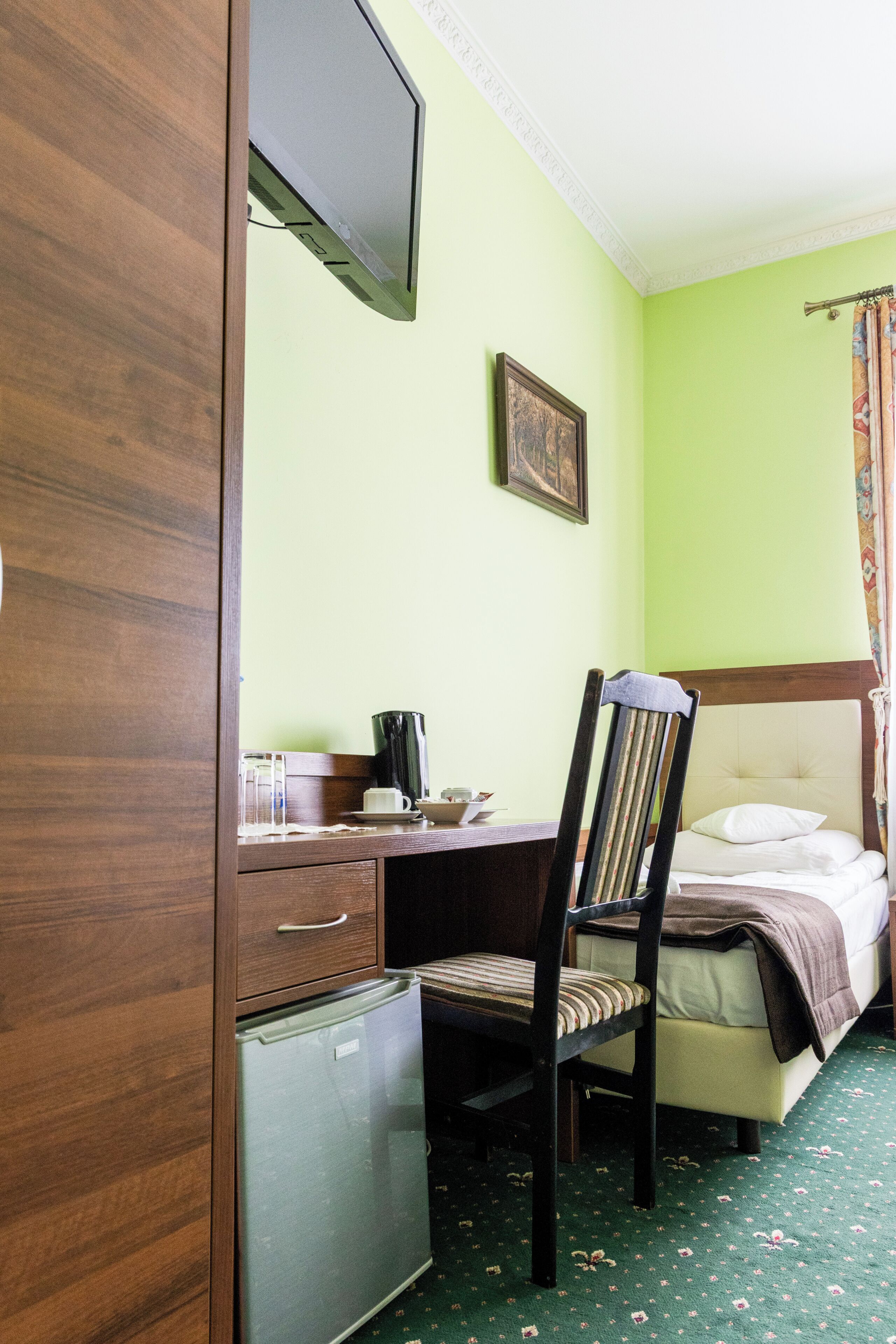 Una camera d'albergo con un letto, una scrivania, una sedia, un piccolo frigorifero e un televisore appeso al muro.