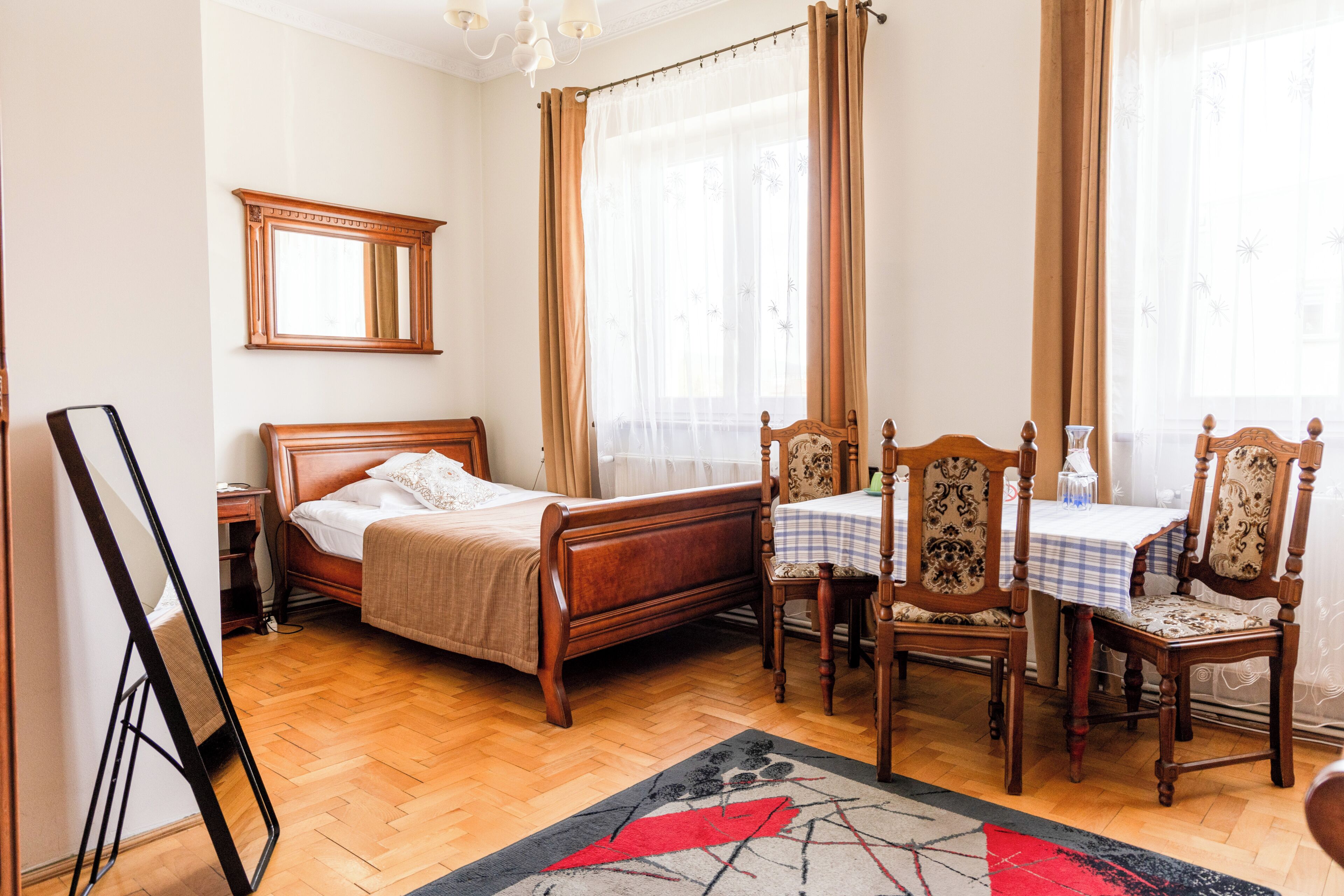 A bedroom with a wooden bed, a bedside table, a chair, a table with a checkered tablecloth, and a mirror on the wall.