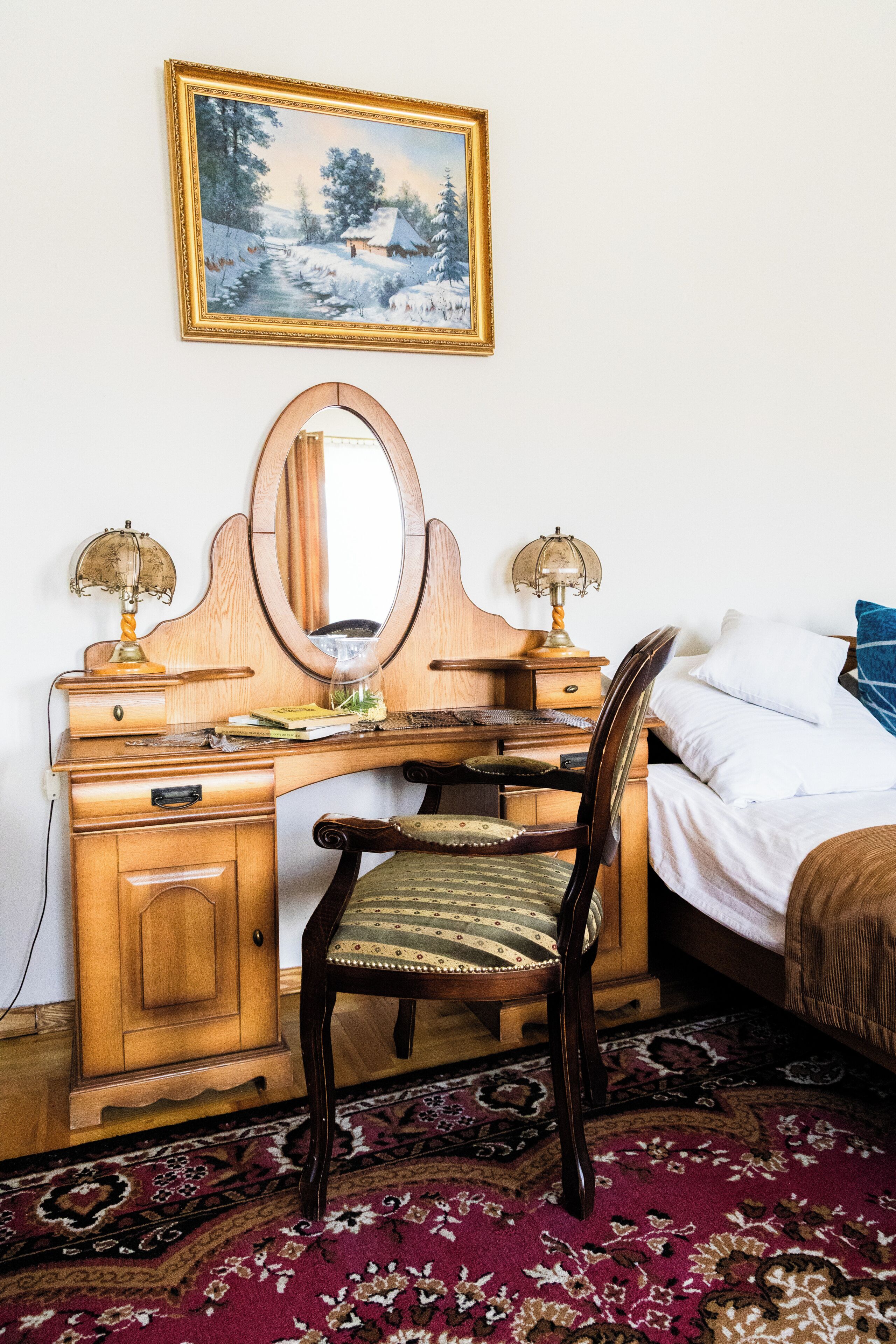 A wooden desk with a mirror, two lamps, and a chair in a room with a bed and a painting on the wall.