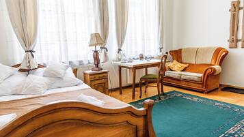 A hotel room with a bed, a wooden nightstand, a table with a lamp, a chair, a sofa, and a patterned rug.